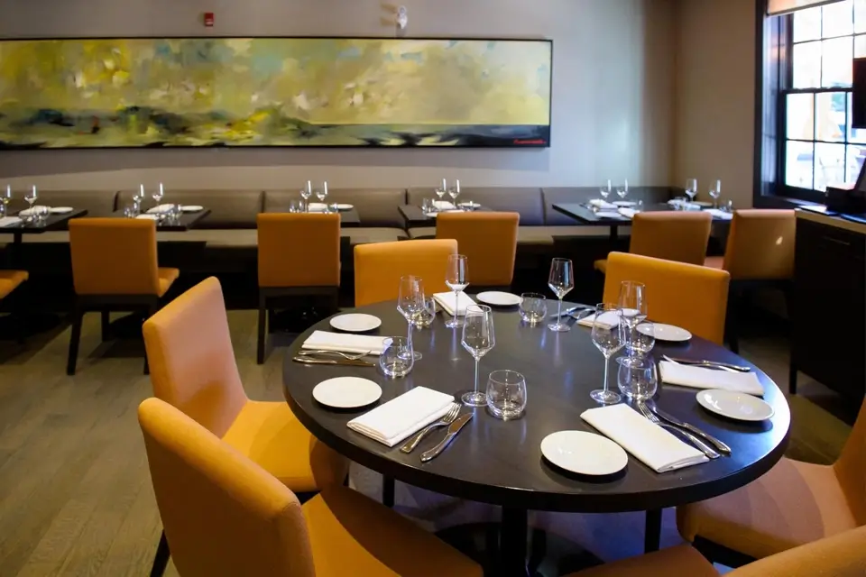 A dimly lit restaurant interior featuring a round table set with white plates, glasses, and silverware. Orange chairs surround the table. A long, abstract painting decorates the wall, and a window to the right lets in natural light. 124 on Queen Hotel and Spa | Hotel & Spa Niagara-on-the-lake | 1 (855) 988-4552