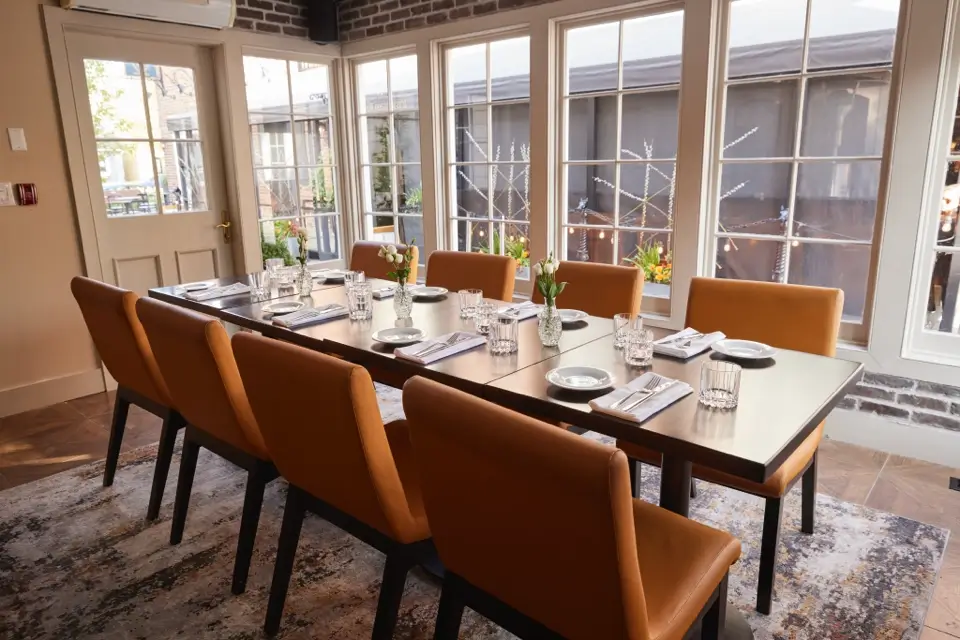 A dining room with a long table set for eight. The table features folded napkins, plates, glasses, and small vases of flowers. The room has orange chairs, a patterned rug, and large windows allowing natural light in. 124 on Queen Hotel and Spa | Hotel & Spa Niagara-on-the-lake | 1 (855) 988-4552