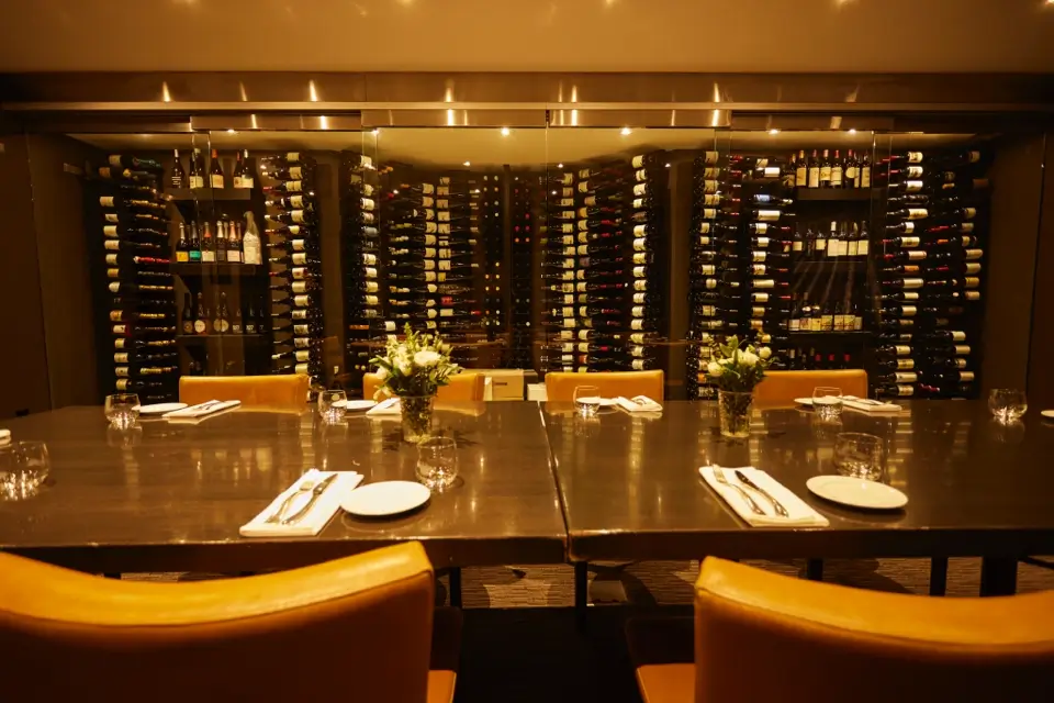 A dimly lit dining room with a long table set for six. The table has yellow chairs, white plates, silverware, glasses, and flower arrangements. The background features a large wine rack filled with bottles. 124 on Queen Hotel and Spa | Hotel & Spa Niagara-on-the-lake | 1 (855) 988-4552