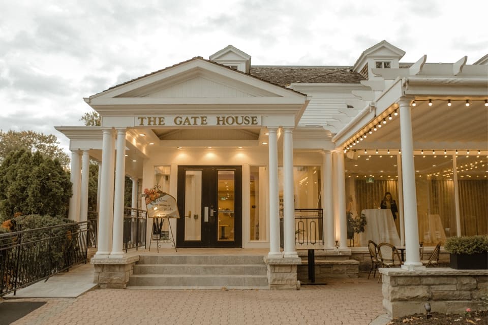 A white building labeled The Gate House exudes elegance as a premier wedding venue, featuring classical columns bathed in warm lighting. The entrance, framed by glass doors on a small stoop, opens under a canopy of string lights stretching to the patio with empty chairs beneath the overcast sky. 124 on Queen Hotel and Spa | Hotel & Spa Niagara-on-the-lake | 1 (855) 988-4552