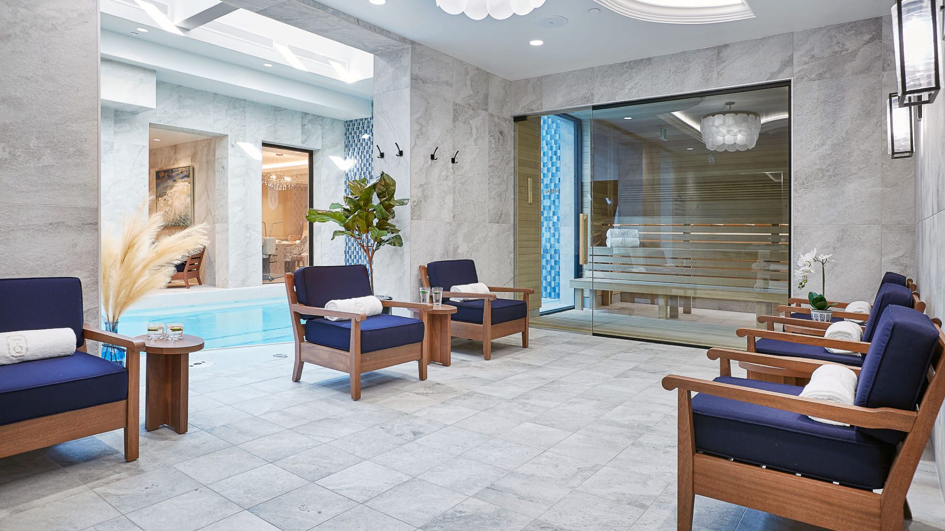 A luxurious spa lounge with blue-cushioned wooden chairs and small tables arranged around a tiled floor. In the background, theres an indoor pool and a glass-walled sauna. Warm lighting and decorative plants create a serene atmosphere. 124 on Queen Hotel and Spa | Hotel & Spa Niagara-on-the-lake | 1 (855) 988-4552