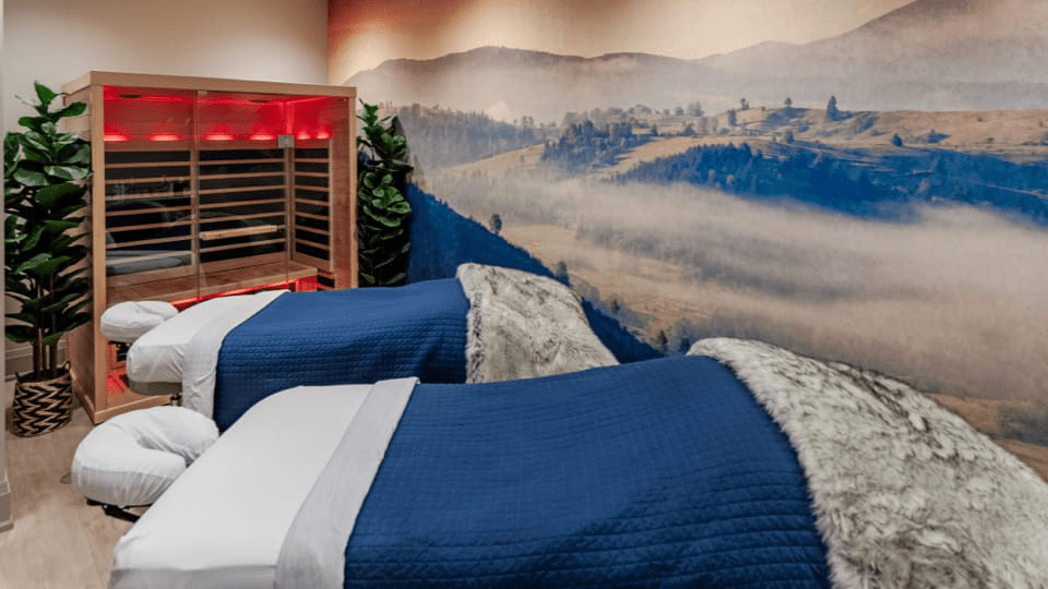 The Spa at Q is your destination spa, featuring a serene room with two massage tables draped in blue blankets and fur throws. A landscape mural of misty mountains graces the wall, while a red-lit wooden sauna in the corner and lush potted plants enhance the calming atmosphere. 124 on Queen Hotel and Spa | Hotel & Spa Niagara-on-the-lake | 1 (855) 988-4552