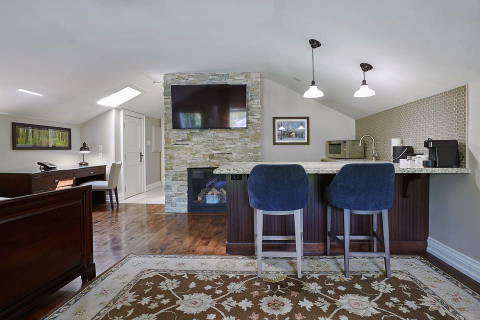 A cozy living space with hardwood floors featuring a stone fireplace, a wall-mounted TV, and a bar area with two blue stools. Theres a desk with a black chair in the corner, and a patterned rug on the floor. Two pendant lights hang above the bar. 124 on Queen Hotel and Spa | Hotel & Spa Niagara-on-the-lake | 1 (855) 988-4552