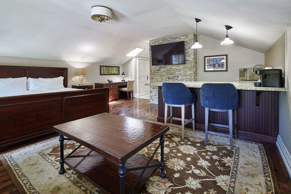 Spacious attic room with a wooden floor, featuring a large bed, a coffee table, a desk, and a TV mounted on a textured stone wall. Two blue bar stools are at a marble counter. A patterned rug adds warmth to the space. Ceiling lights provide illumination. 124 on Queen Hotel and Spa | Hotel & Spa Niagara-on-the-lake | 1 (855) 988-4552