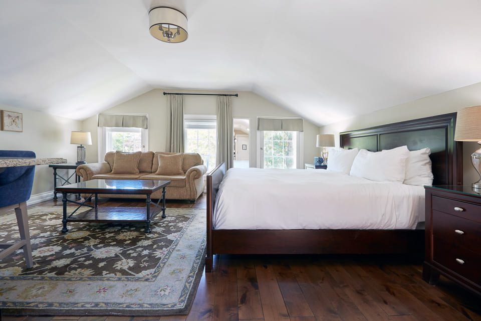 Spacious bedroom with a vaulted ceiling, featuring a large bed with white linens, a sitting area with a beige sofa and glass coffee table, and a gray area rug. Natural light streams through two windows with light curtains and wooden floors enhance warmth. 124 on Queen Hotel and Spa | Hotel & Spa Niagara-on-the-lake | 1 (855) 988-4552