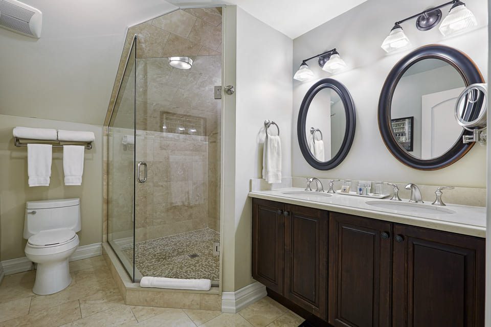 Modern bathroom with a glass-enclosed shower, a toilet with folded towels above, and a dark wood vanity with dual sinks. Two oval mirrors and three-light fixtures are above the vanity, creating a sleek and clean look. 124 on Queen Hotel and Spa | Hotel & Spa Niagara-on-the-lake | 1 (855) 988-4552