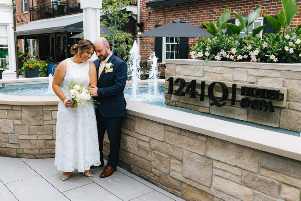 A bride and groom pose affectionately by a stone fountain. The bride holds a bouquet and wears a white lace dress, while the groom is in a dark suit with a boutonniere. The sign reads 1241 QI Hotel & Spa. 124 on Queen Hotel and Spa | Hotel & Spa Niagara-on-the-lake | 1 (855) 988-4552