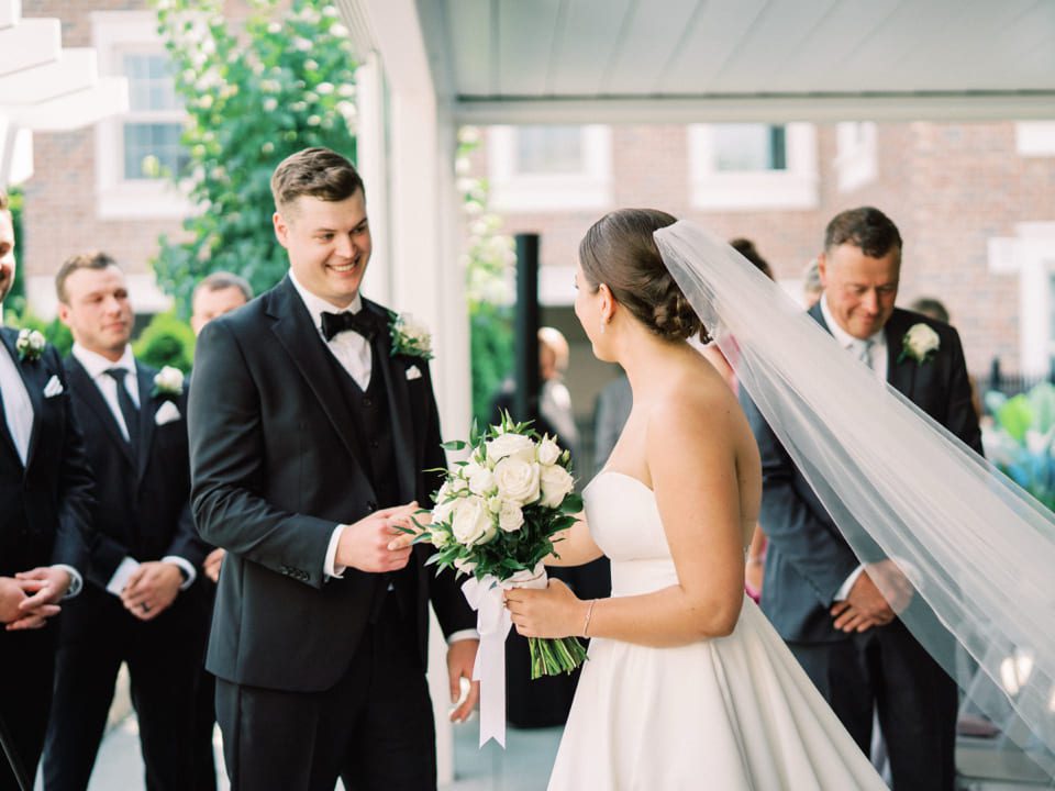 A bride and groom exchange a joyful glance during an outdoor wedding ceremony. The bride holds a bouquet of white roses, her veil flowing. Groomsmen and ushers in black suits stand nearby, smiling, against a backdrop of greenery and a brick building. 124 on Queen Hotel and Spa | Hotel & Spa Niagara-on-the-lake | 1 (855) 988-4552