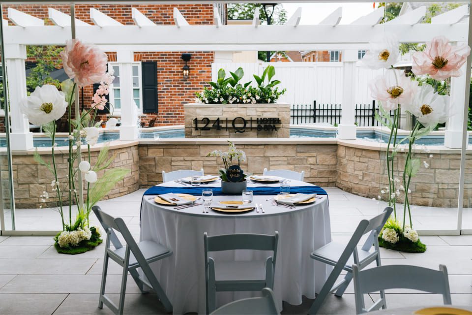 Round table set for four with white chairs and a navy blue runner under a pergola. Large artificial flowers flank the table with a small pond and 124 on Queen Hotel sign in the background. 124 on Queen Hotel and Spa | Hotel & Spa Niagara-on-the-lake | 1 (855) 988-4552