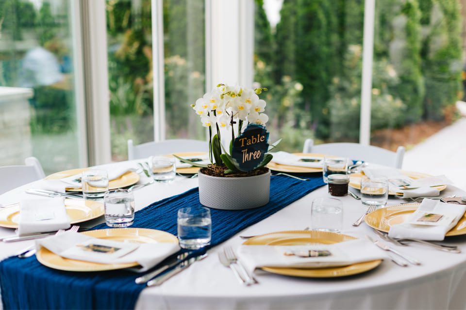A round table is elegantly set with a blue table runner, gold-rimmed plates, and white napkins. A centerpiece features white flowers in a pot marked Table Three. The table is surrounded by large windows with a green garden view. 124 on Queen Hotel and Spa | Hotel & Spa Niagara-on-the-lake | 1 (855) 988-4552