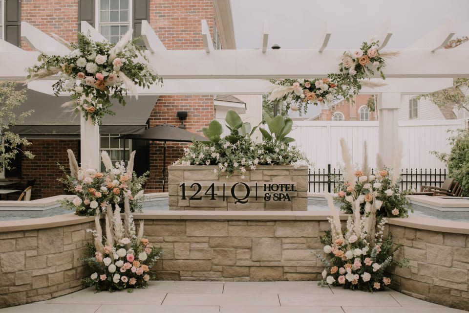 Beautiful outdoor area of a hotel and spa, featuring elegant floral arrangements with pink and white flowers. A pergola and brick wall frame the scene, creating a serene atmosphere. The sign reads 1241 Q Hotel & Spa. 124 on Queen Hotel and Spa | Hotel & Spa Niagara-on-the-lake | 1 (855) 988-4552