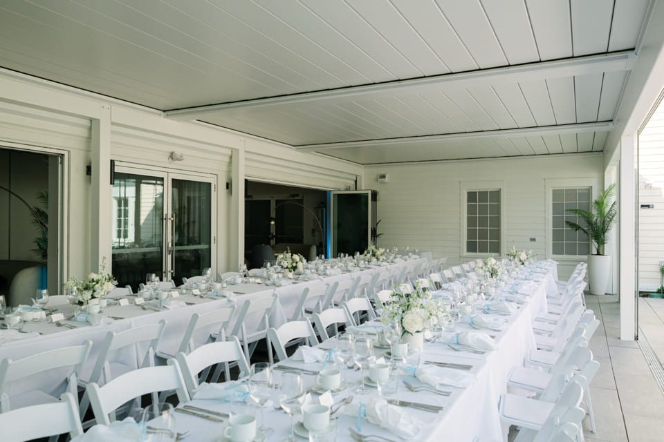 A long dining table is elegantly set for a formal event on a covered patio. White chairs and tablecloths complement the neatly arranged dinnerware and flower centerpieces. The space is enclosed with white walls and large glass doors. 124 on Queen Hotel and Spa | Hotel & Spa Niagara-on-the-lake | 1 (855) 988-4552