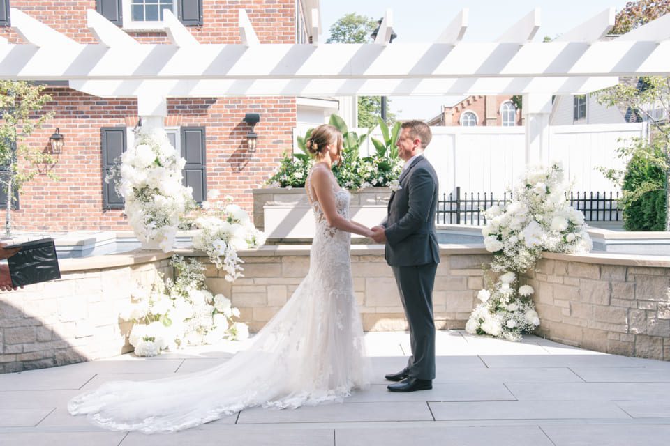 A bride and groom stand facing each other under a pergola in an outdoor setting. The bride is in a lace gown with a long train, and the groom is in a suit. They are surrounded by white floral arrangements. The background features brick walls and white fencing. 124 on Queen Hotel and Spa | Hotel & Spa Niagara-on-the-lake | 1 (855) 988-4552