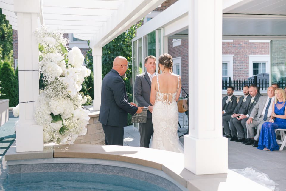A couple stands together under a white pergola during an outdoor wedding ceremony. A man officiates the ceremony. Guests are seated nearby. The bride wears a backless white gown with floral lace details, and the groom is in a gray suit. White flowers decorate the scene. 124 on Queen Hotel and Spa | Hotel & Spa Niagara-on-the-lake | 1 (855) 988-4552