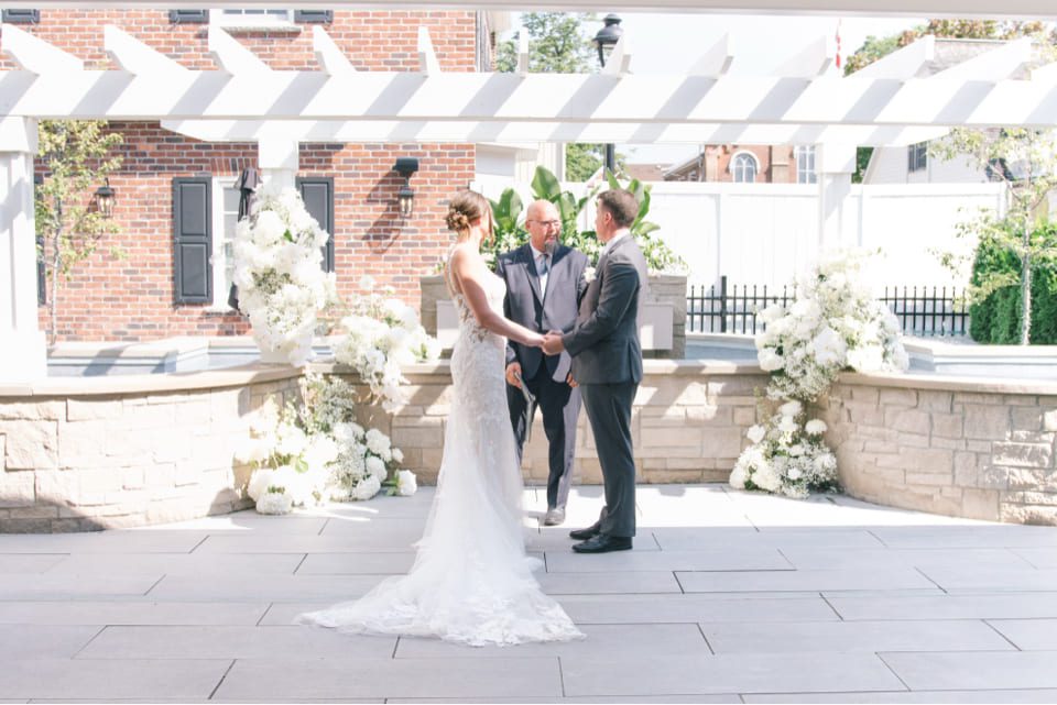 A bride and groom stand facing each other, holding hands, under a pergola with white flowers and greenery. A person stands behind them officiating the ceremony. The setting is an outdoor patio with brick and stone elements. 124 on Queen Hotel and Spa | Hotel & Spa Niagara-on-the-lake | 1 (855) 988-4552