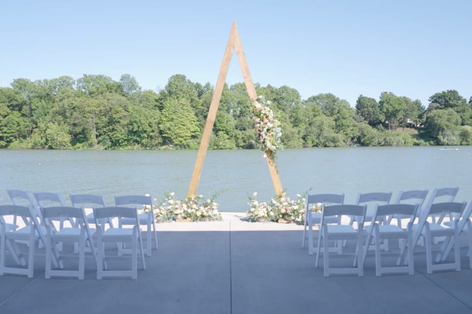An outdoor wedding ceremony setup by a lake, featuring white chairs in rows facing a wooden triangular arch decorated with flowers, with trees and water in the background under a clear sky. 124 on Queen Hotel and Spa | Hotel & Spa Niagara-on-the-lake | 1 (855) 988-4552