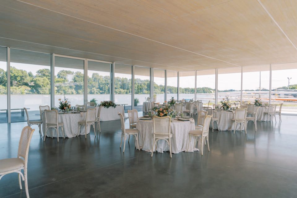 A bright, modern event space with large windows overlooking a river. Round tables with white tablecloths and floral centerpieces are arranged around the room, with white chairs at each table. Trees are visible outside. 124 on Queen Hotel and Spa | Hotel & Spa Niagara-on-the-lake | 1 (855) 988-4552
