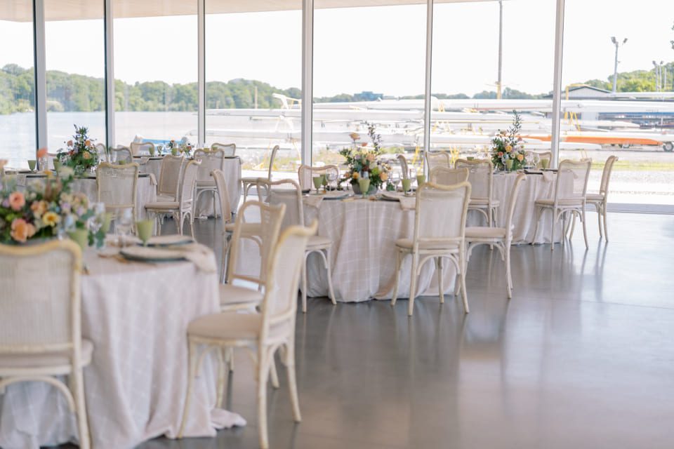 Elegant indoor event space with round tables covered in white linens, decorated with floral centerpieces and surrounded by white chairs. Large windows reveal a view of airplanes and greenery outside. 124 on Queen Hotel and Spa | Hotel & Spa Niagara-on-the-lake | 1 (855) 988-4552