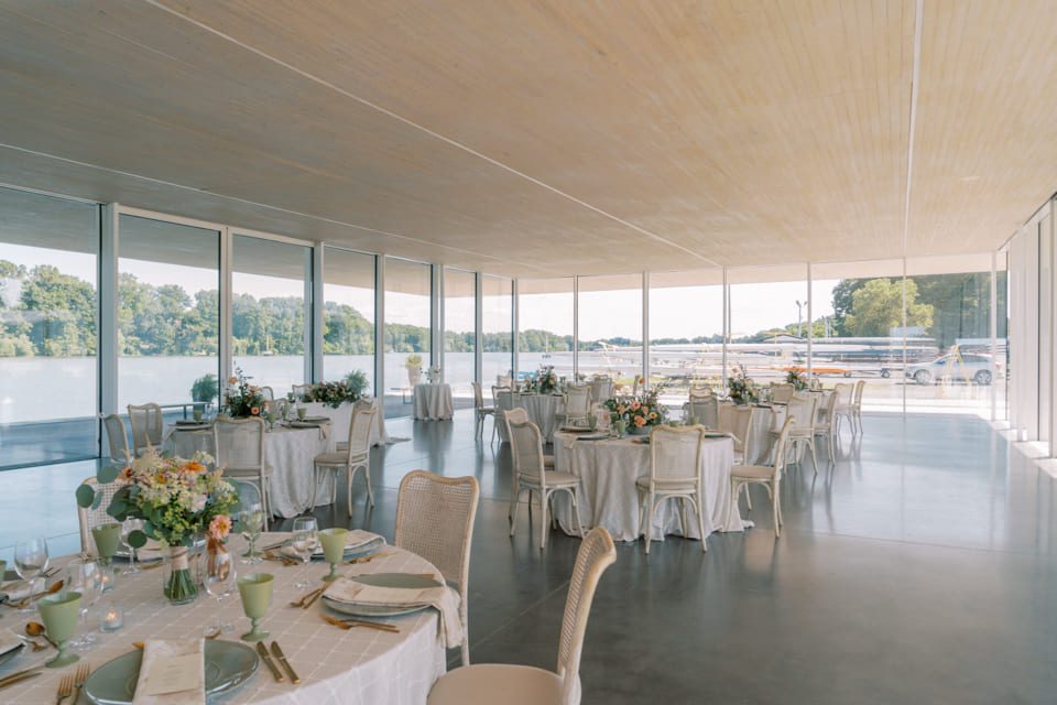 A modern, bright event space with large floor-to-ceiling windows overlooking a lake. Several round tables are elegantly set for a formal gathering, decorated with floral centerpieces and white linens. 124 on Queen Hotel and Spa | Hotel & Spa Niagara-on-the-lake | 1 (855) 988-4552