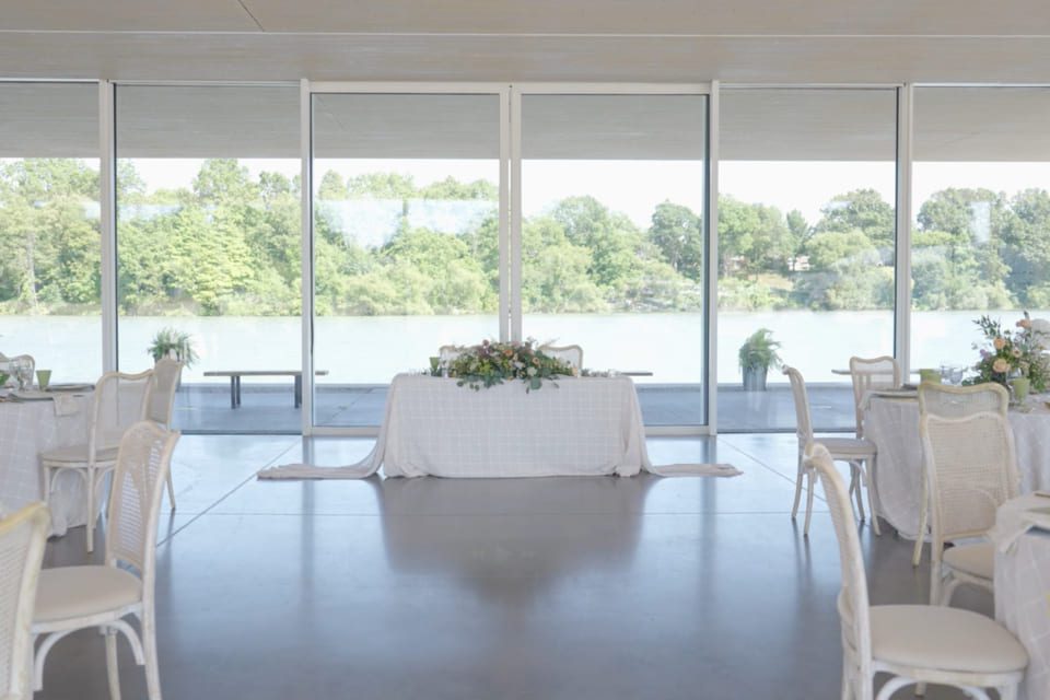 A bright, elegant indoor event space with white tables and chairs arranged around a central table. Large glass windows reveal a scenic lake and green trees outside. Floral decorations are placed on the tables. 124 on Queen Hotel and Spa | Hotel & Spa Niagara-on-the-lake | 1 (855) 988-4552