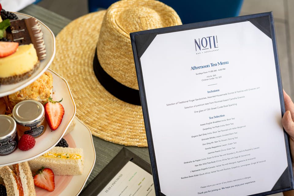 A hand holds an afternoon tea menu next to a two-tiered tray with desserts, scones, fruit, and sandwich slices. A straw hat sits in the background on the table. 124 on Queen Hotel and Spa | Hotel & Spa Niagara-on-the-lake | 1 (855) 988-4552