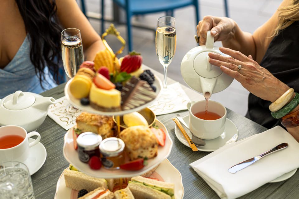 Two women enjoy afternoon tea with a tiered tray of desserts, scones, and sandwiches. One pours tea into a cup, and there are glasses of sparkling wine on the table. 124 on Queen Hotel and Spa | Hotel & Spa Niagara-on-the-lake | 1 (855) 988-4552