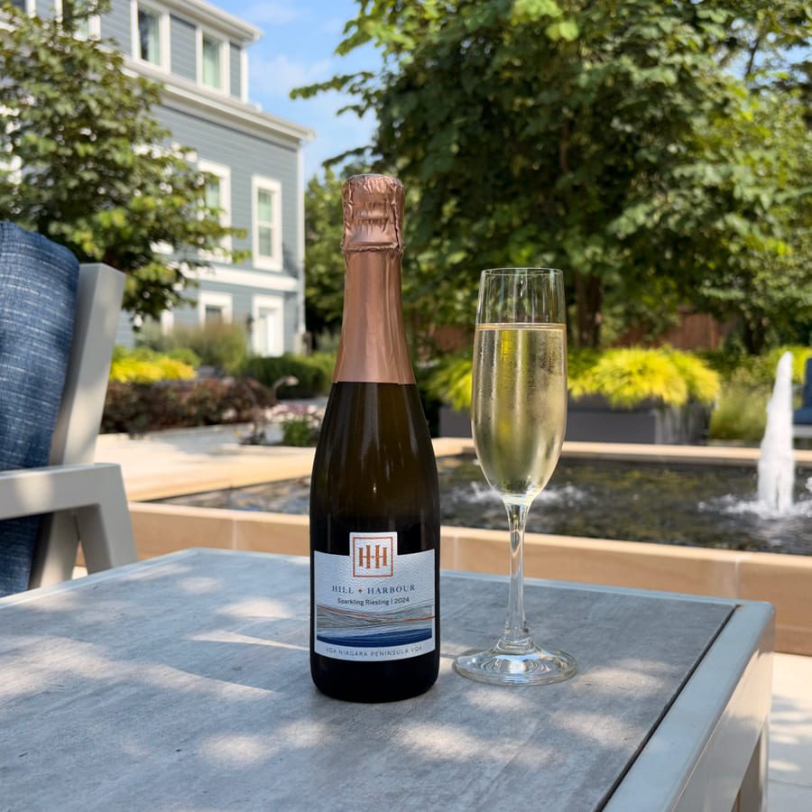 A bottle of sparkling wine and a filled champagne flute sit on a patio table outdoors, with a fountain, trees, and a house in the background on a sunny day. 124 on Queen Hotel and Spa | Hotel & Spa Niagara-on-the-lake | 1 (855) 988-4552