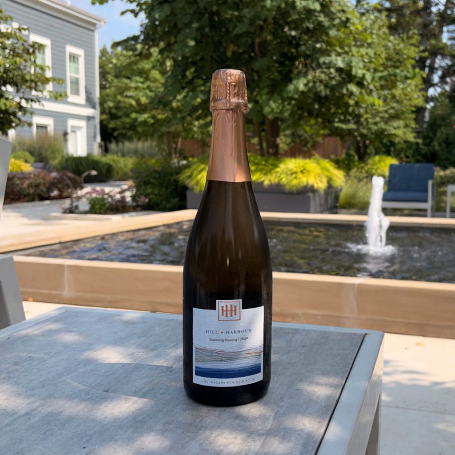 A bottle of sparkling wine sits on an outdoor table, with a small fountain, greenery, and part of a house visible in the background on a sunny day. 124 on Queen Hotel and Spa | Hotel & Spa Niagara-on-the-lake | 1 (855) 988-4552