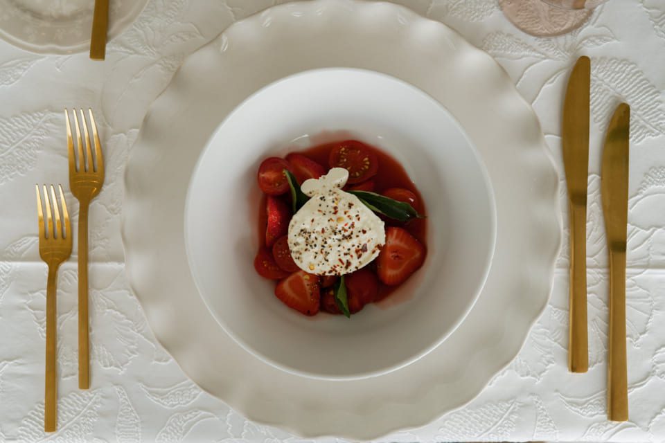 A white bowl with burrata cheese, tomatoes, and basil, placed on a white textured tablecloth. Gold-colored fork, knife, and spoon are arranged on either side of the plate. 124 on Queen Hotel and Spa | Hotel & Spa Niagara-on-the-lake | 1 (855) 988-4552