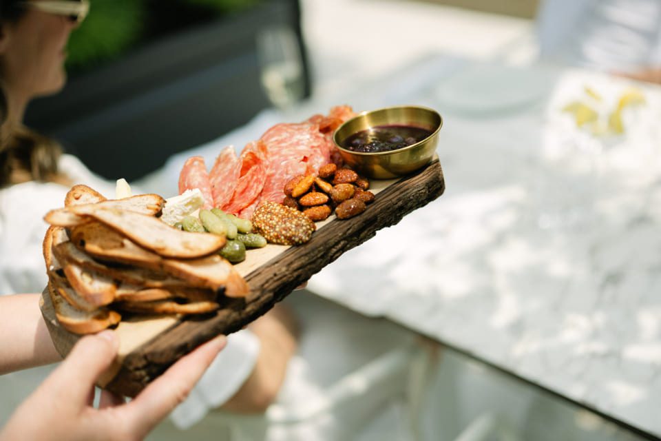 A person holds a rustic wooden board featuring a charcuterie spread with sliced baguette, salami, cornichons, coarse mustard, roasted nuts, and a small bowl of dark jam or spread. The setting appears to be an outdoor lunch table. 124 on Queen Hotel and Spa | Hotel & Spa Niagara-on-the-lake | 1 (855) 988-4552